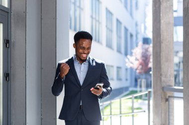 Smartphone 'a bakarak başarıyı kutlayan Afrikalı Amerikalı iş adamı çıktı. Başarılı bir iş adamı zaferi dışarıda cep telefonuyla kazanır. Mutlu İyi Haberler Başarı Anlaşması, Başlangıç Bahis Kazanma oranı