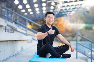 Başarılı bir sporcu sabah meditasyon yaparken lotus pozisyonunda oturur, Asyalı bir adam kameraya bakar ve baş parmağını kaldırarak gülümser.