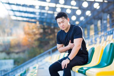 Erkek Asyalı koşucunun göğüs ağrısı var. Koşu ve antrenmandan sonra kalp ağrısı çekiyor. Stadyumun dışında.