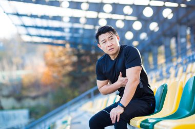 Erkek Asyalı koşucunun göğüs ağrısı var. Koşu ve antrenmandan sonra kalp ağrısı çekiyor. Stadyumun dışında.