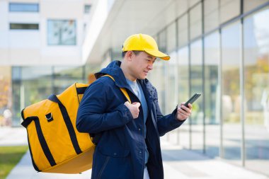 Asyalı erkek yemek kuryesi cep telefonu uygulamasını akıllı telefondan kullanırken termal sırt çantasıyla izole bir şekilde yemek dağıtımı yapıyor. Müşteri adresine bakıyorum. çevrimiçi harita al