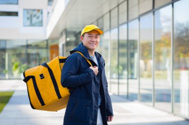 Yürüyen kurye Asyalı gülümseyen ve kameraya bakan yiyecek teslimatı için büyük sarı bir sırt çantası var.
