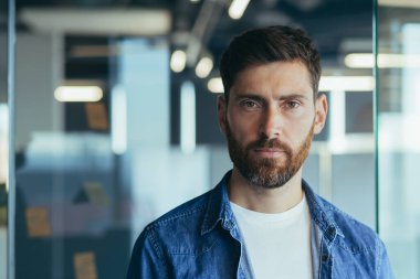 Yeni başlayan, sakallı, iş adamı, ciddi düşünen bir liderin portresi modern ofiste gündelik kıyafetlerle kameraya yakından bakıyor.