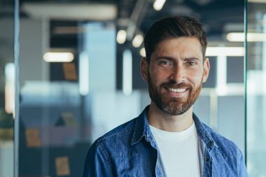 Başarılı bir iş adamı, sakallı, gülümseyen ve kameraya bakan mutlu, yakın plan bir fotoğraf, modern ofiste.