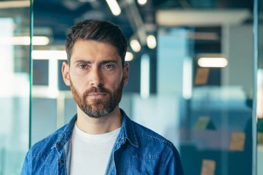 Yeni başlayan, sakallı, iş adamı, ciddi düşünen bir liderin portresi modern ofiste gündelik kıyafetlerle kameraya yakından bakıyor.