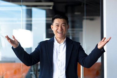 Asyalı mutlu adam kameraya bakıyor. Başparmak işareti yapıyor. Modern ofiste gülümseyen bir işadamı.