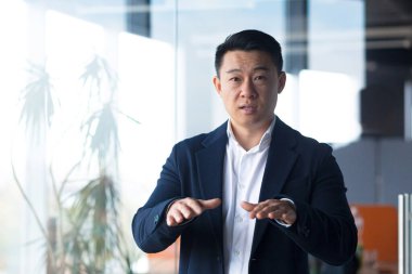 Ciddi Asyalı işadamı kameraya bakar çevrimiçi bir toplantı düzenler, el hareketleriyle bir adam görevi meslektaşlarına açıklar.