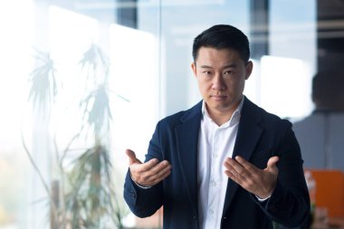 Ciddi Asyalı işadamı kameraya bakar çevrimiçi bir toplantı düzenler, el hareketleriyle bir adam görevi meslektaşlarına açıklar.