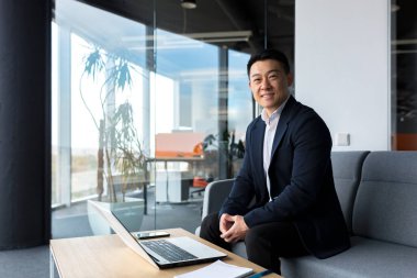 Başarılı ve mutlu Asyalı adam online olarak dizüstü bilgisayarla çalışıyor. Plan ve görevle mutlu bir şekilde gülümsüyor.