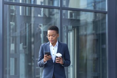Kendine güvenen bir Afro-Amerikalı kadın iş bölgesinde dolaşıyor ve telefonu kullanıyor. Elinde bir fincan kahve, resmi giysiler içinde. İletişim kurarken modern akıllı telefonlara göz atan çekici bir iş kadını. uygulamalar
