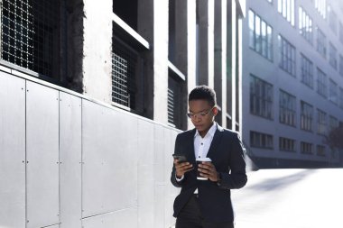 Ciddi kız öğrenci düşünme, elinde telefon tutma ve kullanma, Afro-Amerikan iş kadını elinde bir bardak sıcak içecekle ofisin yakınında yürüyor.