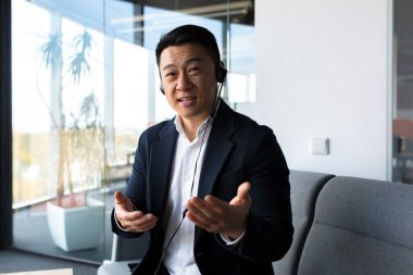 Asyalı dost canlısı web kamerasına bakıyor ve konuşuyor, kulaklıklı işadamı araya giren kişiye gülümsüyor, modern ofisten video çağrısı