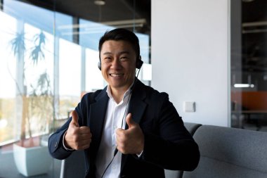 Asyalı dost canlısı web kamerasına bakıyor ve konuşuyor, kulaklıklı işadamı araya giren kişiye gülümsüyor, modern ofisten video çağrısı