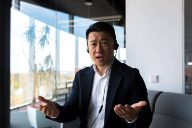 Sorusu olan bir adam kameraya bakar, Asyalı bir işadamı kulaklıkla video görüşmesi yapar.