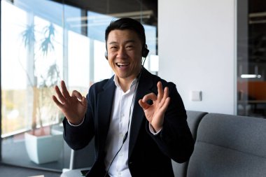 Asyalı dost canlısı web kamerasına bakıyor ve konuşuyor, kulaklıklı işadamı araya giren kişiye gülümsüyor, modern ofisten video çağrısı