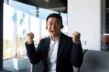 Asyalı dostça webcam ve konuşmalara bakıyor, kulaklıklı işadamı araya giren kişiye gülümsüyor, modern ofisten gelen video zaferi kutluyor