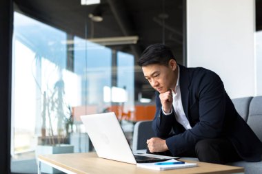 Modern bir ofis masasında dizüstü bilgisayarda çalışan düşünceli Asyalı iş adamı portresi. Kendine güveni tam, resmi takım elbiseli dalgın bir adam. Bir sorunu çözmek için ilham almayı düşünüyorum. Açılış