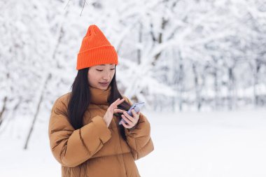 Parkta yürüyen güzel Asyalı kadın, kar yağışlı bir günde internetten alışveriş için telefonu kullanıyor.