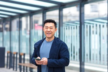 Cheerful and happy asian using phone smiling and looking at camera, near airport, businessman on tourist trip