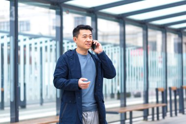 A man talking on the phone, focused and serious Asian at a public transport stop, a businessman near the office center