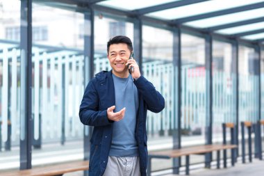 Cheerful and happy Asian businessman talking on the phone, waiting for a plane and a taxi at a public transport stop