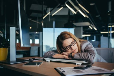 Beautiful blonde business woman sleeping at the table, at her workplace at the computer, in a modern office, working late, overworked