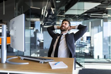 Happy and dreamy businessman, resting at work with his hands behind his head, working in a modern office at the computer