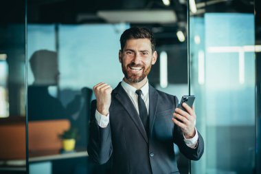 Happy businessman boss looks at camera and smiles, holds phone in hands, man wins online and celebrates victory