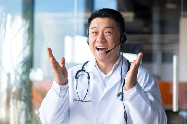 快乐的医生微笑着看着摄像机 亚洲人告诉病人好消息 用视频通话 用耳机和同事们在网上见面 用手拿着有趣的手势 用网络摄像头观看 — 图库照片
