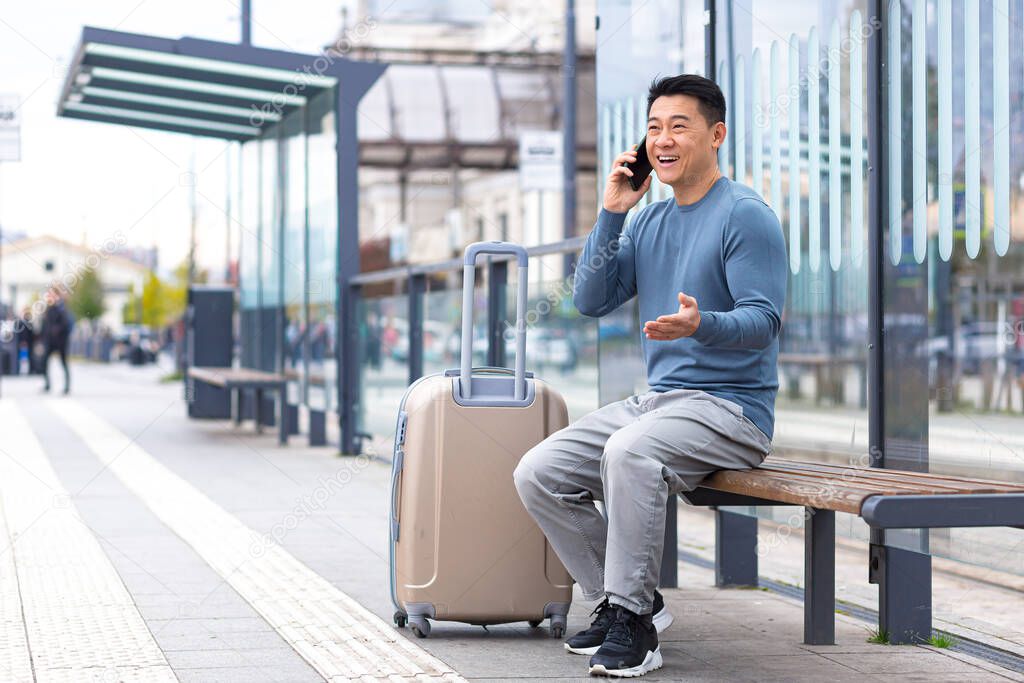 Tourist at the bus station, having fun talking on the phone, sitting on a bench with a large suitcase waiting for a taxi, Asian man on a business trip