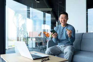 Happy asian male businessman holding credit card and phone, online shopping worker sitting in office