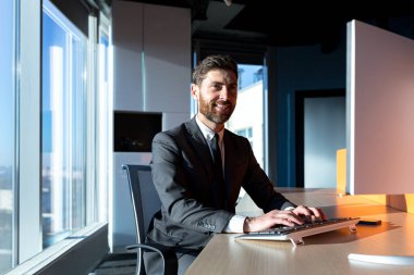 Happy successful businessman with a beard working at a computer in a modern office, smiling and rejoicing, confident boss