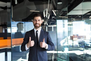 Happy and successful businessman looking at camera and smiling, approvingly showing thumbs up in modern office