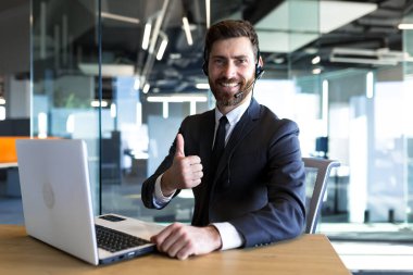 Happy and successful call center employee looks at the camera and smiles, businessman keeps his finger approvingly up, working at the computer, uses a headset for a video call