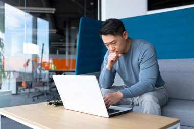 A thinking Asian man works at a computer in a modern office, a freelancer is focused on solving a task, makes an important job.