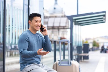 Cheerful and happy Asian businessman talking on the phone, waiting for a plane and a taxi at a public transport stop