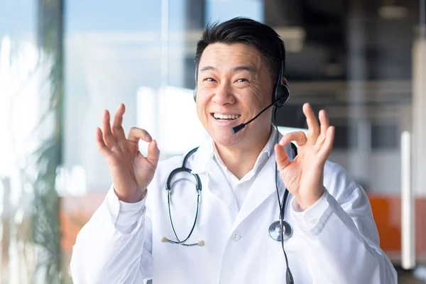 一个经验丰富的亚洲医生的画像 看着摄像机 与病人愉快地交谈 友善地微笑 为病人的康复感到高兴 远程工作 带耳机的视频通话 — 图库照片