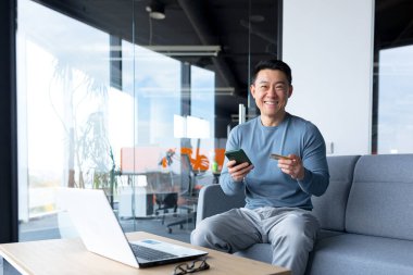 Successful and happy Asian makes online purchases with a credit card and an application on the phone, a businessman spends time in a modern office on a laptop
