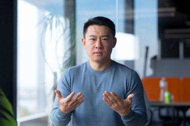 Portrait of a serious and angry employee, looking at the camera and gesturing with his hands, Asian communicates on a video call, looks at the webcam