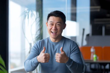 Portrait of a cheerful employee, looking at the camera and gesturing with his hands, Asian communicates on a video call, looks at the webcam