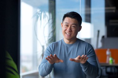 Portrait of a cheerful employee, looking at the camera and gesturing with his hands, Asian communicates on a video call, looks at the webcam