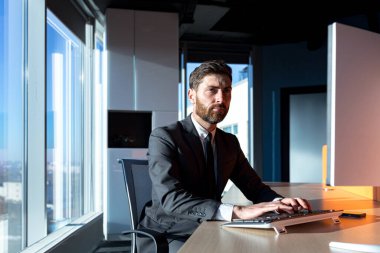 Focused broker working at computer in marvelous office by the window, male businessman in business suit