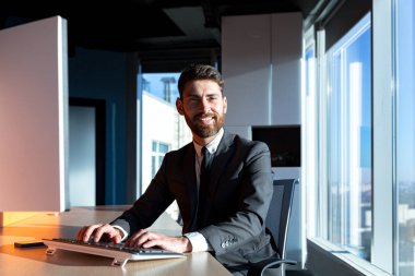 Happy and smiling businessman working in modern office by the window at the computer, Bearded boss owner