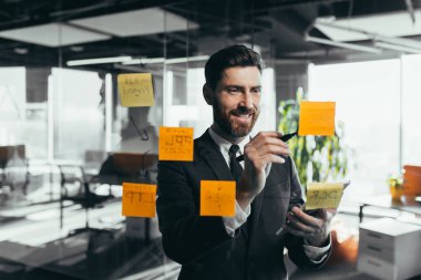 Successful businessman rewrites a task on a glass board with colored stickers, from phone and tablet