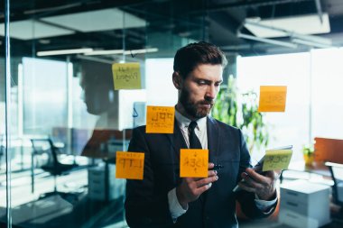 Successful businessman rewrites a task on a glass board with colored stickers, from phone and tablet
