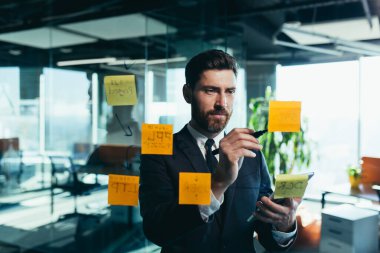 Successful businessman rewrites a task on a glass board with colored stickers, from phone and tablet