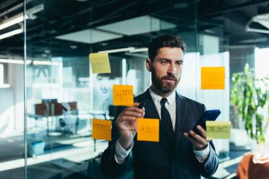 Successful businessman rewrites a task on a glass board with colored stickers, from phone and tablet