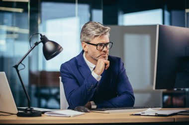 A gray-haired man with glasses thinks about the tasks, a businessman with a beard works in a modern office at the computer,