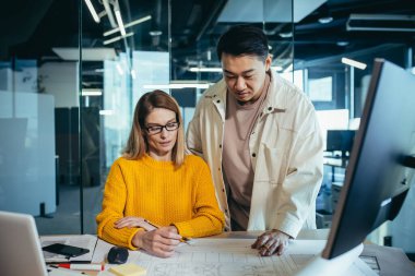Tasarımcı ve mimarların iki çalışanı, projeyi düşünüyorlar, Asyalı bir adam ve sarışın bir kadın, gündelik giysiler içinde masada birlikte çalışıyorlar, bir plan çizimi projesiyle birlikte.