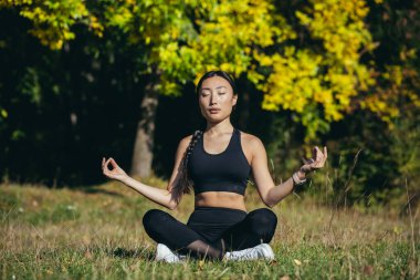 Young beautiful asian woman performs fitness yoga exercises, sitting in lotus position on the grass and meditating in the park